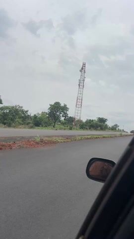 The video starts with a shot from inside a moving vehicle, showing a tall, red-and-white communication tower along a rural road. The camera then shifts to focus on a man in the front seat of the car, who is talking to the camera. A second person can be seen in the background, seated behind him. The scene is overcast. The man has dark skin, a beard, and is wearing a black shirt with a gold necklace. The car is moving along an asphalt road with sparse vegetation on the roadside.