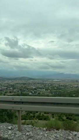 The video starts with a view of a cloudy sky and distant town, then focuses to a car traveling on a mountain road with a view of the landscape. Other cars pass in the opposite direction. The video seems to be filmed from the dashboard of a car.