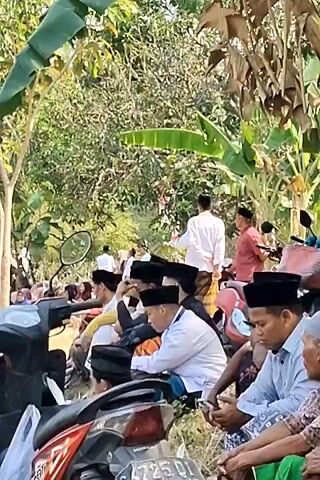 The video shows a gathering of people outdoors, many of whom are wearing traditional Islamic attire, including peci caps. The scene appears to be a religious or cultural event. In the foreground, several men are seated on the ground, some are looking at their phones, while others are conversing. Behind them, more people are visible, some sitting and others standing. Further in the background, there is a tent set up, under which a group of people are seated, possibly for prayer or a ceremony. There are also trees and vegetation surrounding the area, suggesting an outdoor rural setting. Several motorcycles are parked nearby, indicating that attendees likely traveled by this mode of transport. Red and white flags, possibly national flags, are also visible, adding to the sense of a community event. The overall atmosphere seems peaceful and communal.