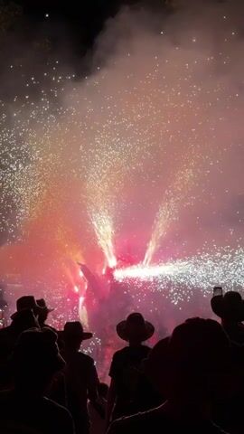 The video shows a crowd of people silhouetted against a pyrotechnic display. The fireworks are a vibrant mix of red and orange, with sparks and plumes of smoke filling the night sky. The people in the foreground are wearing hats and appear to be looking up at the fireworks, with some holding up their phones to record the event. The overall impression is one of a festive outdoor gathering.