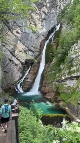Here is a description of the video:

The video shows a tall, thin waterfall cascading down a rocky mountainside. The water falls into a clear, turquoise pool. Lush green vegetation clings to the rocks surrounding the waterfall. A wooden fence runs along a path in the foreground, where several people are standing and observing the scene. The overall atmosphere is one of natural beauty and tranquility.
