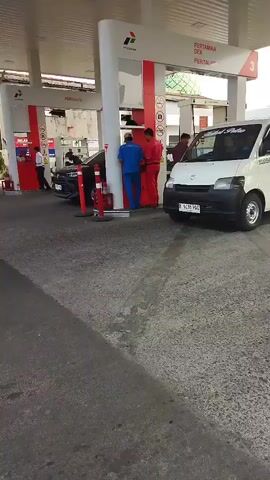 This video shows a gas station with multiple vehicles and employees. In the foreground, the asphalt pavement is visible. In the background, there are several gas pumps under a white canopy with red accents. A white van is parked at one of the pumps. A black car is also at a pump, with its hood open, and two male employees, one in a blue uniform and the other in a red uniform, are interacting with the car. Another employee in a white shirt is visible near another pump. A person in a white shirt and black pants is standing by a black car in the background. The environment appears to be an outdoor urban setting.