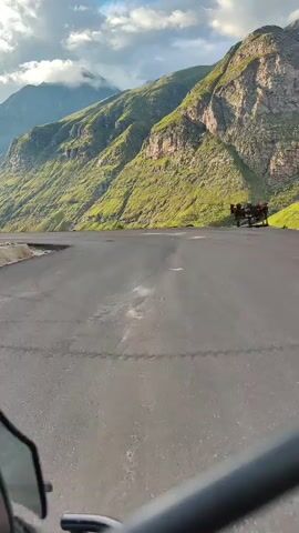 The video shows a point-of-view shot from a vehicle driving on a paved mountain road. The road curves to the left, revealing a steep, green, rocky mountainside on the right. In the distance, more mountains and a cloudy sky are visible. The road appears to be in good condition, with a few scattered puddles. There is some slight wear and tear on the road surface, with some small potholes visible. The vehicle appears to be moving at a moderate speed. The overall impression is of a scenic drive through a mountainous region.