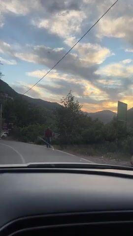 The video shows a car ride through a mountainous area, likely during sunset. The video begins with a man walking on the side of a road. The camera then pans up to show the sunset over the mountains. The view is partially obstructed by trees and power lines. As the car drives, the camera shows the side view mirror and the sky. The car passes buildings and parked vehicles on the side of the road. The setting is bright and colorful due to the sunset.