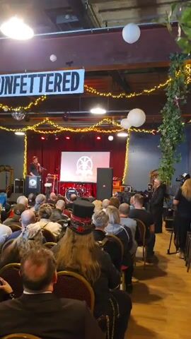 The video shows a large indoor gathering. A speaker is on stage, behind a podium, talking to a seated audience. The speaker is wearing a suit and using a microphone. A screen behind the speaker displays a logo. The room is decorated with string lights and white paper lanterns. A banner hangs from the ceiling that reads "LET US STAND UNBOWED and UNFETTERED." The audience is diverse, with people of various ages and clothing styles. Some audience members are taking photos. The room appears to be a repurposed industrial space with exposed ceilings and brick walls. There are speakers on either side of the stage and mirrors along the side walls.