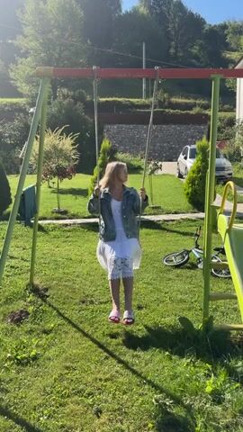 A young woman with blonde hair is swinging on a swing set in a grassy yard. She is wearing a white dress and a denim jacket, with pink slides on her feet. The swing set is green and red, and there is a slide to the right of the frame. In the background, there are trees, bushes, a wall, and two cars parked on a driveway. The sun is shining, casting shadows on the grass. The woman is smiling and looking up.