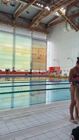 A wide shot captures an indoor swimming pool with multiple lanes demarcated by lane ropes. On the right side of the frame, a woman wearing a colorful swimsuit stands and looks at her phone. In the background, several people are visible near the pool edge. A large digital scoreboard displays text and numbers, reading "16-10-2023 9:46:32". The pool area is brightly lit by overhead lights, and large windows with scenic murals are visible along the far wall. The architecture of the ceiling features exposed wooden beams. The foreground is occupied by the tiled floor surrounding the pool. In the final seconds of the clip, a medium shot focuses on a man with a shaved head, wearing blue swimming briefs, who is standing by the pool.