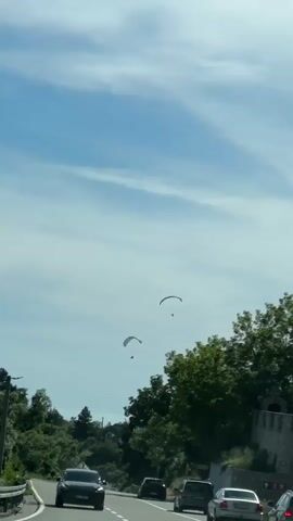 The video shows a road with cars driving. In the sky above, there are paragliders. The sky is blue with some clouds. There are trees on the sides of the road.