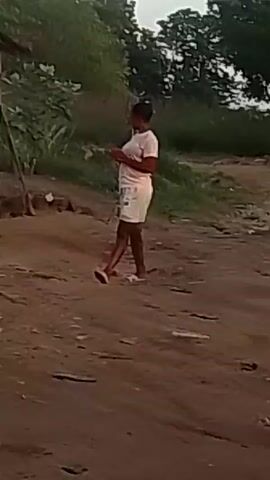 The video shows a young person standing in a dusty area with some buildings in the background. The person is wearing a white t-shirt, white shorts, and white sandals. They are dark-skinned and have short hair. They bring their hands together in front of their chest and appear to be practicing a karate-type movement. The background includes a wooden structure with a corrugated metal roof, greenery, and a few scattered objects like buckets and a pole. The scene is shot in daylight.