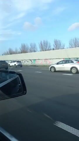 The video shows a view from the driver's seat of a vehicle moving on a highway. Another grey SUV passes to the right. The right side mirror of the recording vehicle is visible in the lower left corner of the frame. Behind a concrete barrier, bare trees are visible against a blue sky with some clouds. Several other cars are present on the road.