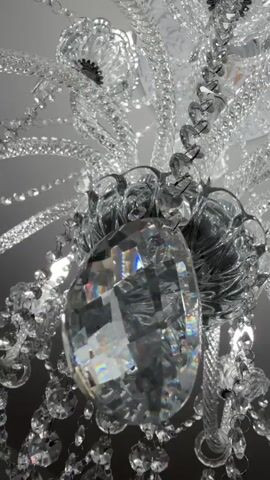 A close-up video shows a crystal chandelier hanging from the ceiling. The chandelier is ornate, with many crystal pendants and strands of beads. The camera moves slowly, offering different perspectives of the chandelier's intricate details. The lighting is dim, with the chandelier's own lights providing illumination, creating a sparkling effect on the crystals. The background is blurred, indicating the focus is solely on the chandelier. The video captures the reflective and dazzling nature of the crystal chandelier.