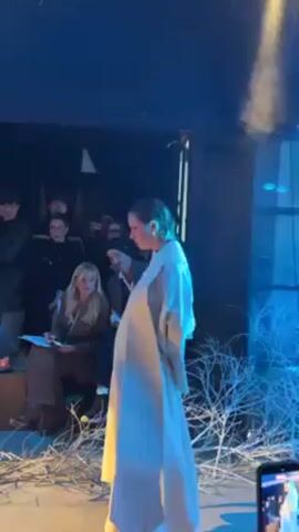 A runway show unfolds in a dimly lit space with blue lighting. A model strides down the runway, wearing a long, off-white coat and orange boots. A seated audience watches and takes notes. The runway is decorated with sparse, white branches. Additional spectators stand along the back of the space and on a balcony above. The venue has visible wooden beams and is decorated with vines. Electronic music plays.