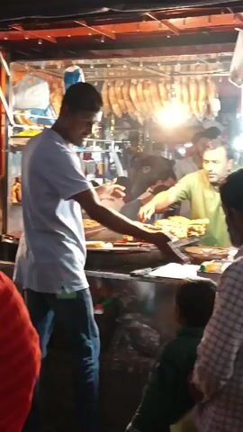 The video depicts a bustling street scene with several people walking around. In the foreground, there's a person wearing a red garment. Behind them, a man in a light-colored shirt is also walking. Further back, there's a food stall with a man preparing and serving food. The stall has food displayed, and there are some containers and utensils visible. Balloons are visible in the background to the left. The scene seems to be set outdoors, likely in a market or a busy street area. The lighting is somewhat dim, suggesting it might be evening or night.