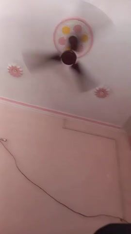 A static shot captures the interior of a room. The ceiling is white with a decorative ceiling fan in the center. The walls are painted a light pink, with a pink decorative border near the ceiling. A door, partially open, is visible in the background, with a purple curtain hanging next to it. A rack with a piece of brown fabric is visible in the lower left. A wire is attached to the wall. The overall scene suggests a simple, lived-in space.