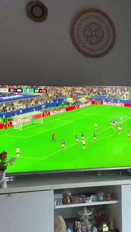 The video shows a television screen displaying a soccer match. The score is 0-0. The game appears to be between France (FRA) and Spain (ESP). The field is green with white lines. There are players in white and blue uniforms on the field. The stands are full of spectators. There are advertisements visible around the field, including McDonald's. The television is on a white cabinet. On the wall above the television, there are two circular decorative items.