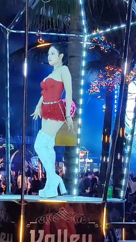 The video shows a woman on a raised platform at night. She is wearing a red strapless top and red sequined shorts, paired with white thigh-high platform boots. Her hair is styled in a bun. The platform is decorated with lights and palm trees. There is a crowd of people visible in the background. The word "Valley" is partially visible on the front of the platform.