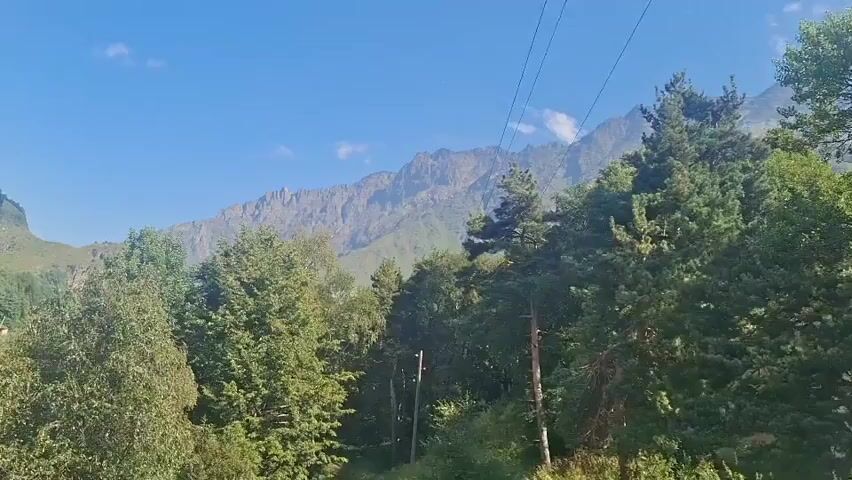 The video displays a panoramic view of a mountainous landscape with a village nestled on a hillside. The camera pans across the scene, revealing lush green trees and a river flowing through the valley. The sky is a clear blue with a few scattered clouds. The village consists of several houses with red roofs, situated on a steep, grassy slope. Power lines and poles are visible throughout the scene, suggesting the presence of infrastructure. The mountains in the background are rugged and imposing, with rocky outcrops and sparse vegetation. The overall impression is one of a remote and picturesque natural setting.