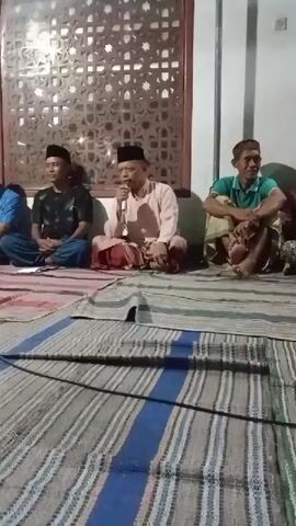 This video shows a group of men sitting on the floor, facing a camera. In the center, a man wearing a pink shirt and red sarong is speaking into a microphone. To his left, a man in a black shirt and to his left, a man in a blue shirt are also seated. To the right of the man with the microphone, a man in a green shirt is seated. In the background, there is a wall with a decorative screen and some lighting. The floor is covered with woven mats. The scene appears to be an indoor gathering, possibly a religious or community event.