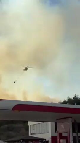 The video shows a firefighting helicopter flying overhead a gas station with a bucket suspended underneath, against a backdrop of smoke-filled skies. The helicopter is positioned in the center of the frame, moving away from the camera with its bucket hanging low. The gas station features a red and white canopy and a sign with a red symbol. A liquid tanker truck is parked near the gas station. In the background, there is a line of trees and a tall electricity tower, all partially obscured by the thick smoke. The sky is overcast with a yellowish tint from the smoke, suggesting a fire nearby.
