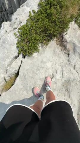 The video begins with a first-person perspective of a person standing on a rocky, mountainous terrain. Their legs, clad in black shorts and pink and white sneakers with white socks, are visible. Green vegetation sprouts from the rocks. The camera then pans up to reveal a vast, scenic landscape of mountains and valleys, covered with dense green forests. The sky is blue with scattered white clouds, and a faint wisp of smoke is visible in the distance. The overall impression is one of a breathtaking natural vista viewed from a high elevation.