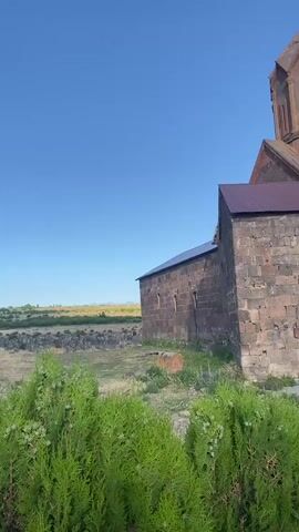 The video shows a panning shot of an ancient stone church with multiple domed towers, set against a clear blue sky. The church is constructed from rough-hewn, dark-colored stones with some lighter stone accents. One of the towers is prominent in the foreground, featuring intricate stonework and a cross at its apex. Another, taller tower is visible in the background, also topped with a cross. The building has a pitched, dark-colored roof. In front of the church, there is a patch of dry, yellowish grass with some scattered rocks. The lower portion of the scene is partially obscured by lush, green foliage, likely bushes or trees. The overall impression is one of historical significance and weathered beauty.