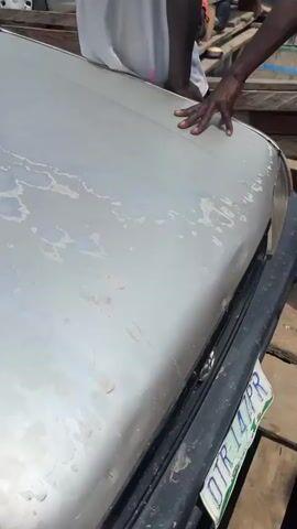 The video begins with a close-up of the hood of a silver car, showing water stains and some damage. A person's hand is visible resting on the hood. The camera then pans to reveal a group of people and several vehicles, including the silver car and a red and black checkered shirted man standing beside it. They are on a wooden structure that appears to be a makeshift bridge or raft over water. In the background, more people and boats are visible on a body of water. The scene suggests a crossing or a ferry operation. The lighting indicates it is daytime. The weather appears to be sunny with clouds in the sky. The overall environment is outdoors and appears to be a rural or less developed area.