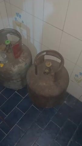 Two gas tanks sit in the corner of a room with blue-tiled flooring and white-tiled walls with flower decorations. The tanks are both rusty and old, one has a pressure regulator attached.  The lighting is dim.