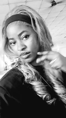 A person with long wavy blonde hair, a black headband, and a dark-colored shirt is the main focus in this black and white video. The person is looking directly at the camera with a neutral expression. They have glossy lips and appear to be wearing jewelry, including a lip ring, a ring on their finger, and a necklace. They briefly raise their hand to their hair. The background is blurry, but there is a glimpse of a ceiling with fixtures.