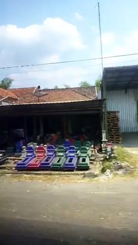 The video shows an outdoor scene. In the foreground, a paved road or pathway is visible. Behind it, several colorful plastic loungers are arranged in a row. These loungers appear to be new and are in various shades of blue, green, and red. To the right of the loungers, there are stacks of wooden pallets and some loose building materials. Further back, a building with a reddish-brown tiled roof is present. A satellite dish is mounted on the roof. The building has an open-fronted structure, possibly a shop or workshop, with various items stored inside. A tall antenna or pole extends upwards from behind the building towards the sky. The sky is blue with some white clouds. The overall environment appears to be rural or semi-urban. There are no people clearly visible in the frame.