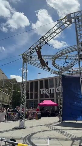 A man in a harness is moving across a high obstacle course. He is wearing khaki clothing and a helmet. The course is made of silver metal scaffolding and suspended cables. There is a crowd of people standing behind a metal barrier watching him. There is a building with "TK Maxx" on its front behind the course and a pink tent. The sky is blue with puffy white clouds. The camera angle is low, looking up at the man on the course.