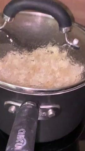 The video shows a covered black saucepan on a black glass cooktop. The saucepan has a clear glass lid with a black handle, and the sides of the pot also have black handles. Inside the saucepan, there appears to be something cooking; it's a mixture of light-colored, chunky material simmering or boiling, causing condensation to form on the underside of the lid. The saucepan is sitting directly on the stovetop. The background shows a plain, light-brown wall. The camera is static, with no zooming or panning. The audio suggests that popcorn is being made in the saucepan.
