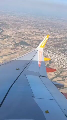The video shows a view from an airplane window. The wing of the plane, with "easyJet" written on it, is prominently in the foreground. Below the wing, a landscape is visible, with fields and some buildings. The sky is hazy and appears to be daytime. There are no people visible in the frame, and no actions or interactions are depicted. The scene is static.