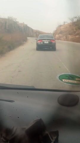 The video shows a view from inside a car driving on a winding road. The road is surrounded by rocky, arid terrain. Ahead, another car is driving in the same direction. The sky is overcast, creating a hazy atmosphere. The dashboard of the car filming the video is visible, with a green and white sticker.