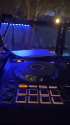 The media file shows a close-up view of a DJ controller with its various buttons, knobs, and jog wheels illuminated by colorful LED lights. The controller is positioned outdoors, with a blurry background that suggests a nighttime setting. Trees and what appears to be a body of water (possibly the sea or a pool) are visible, along with some lounge chairs. The overall ambiance is that of a party or event, likely at a resort or beachside location in Pečurice, Montenegro.

The DJ equipment is the primary focus, with blue and orange lights casting a vibrant glow on its surfaces. The lights on the controller are blinking and changing, suggesting that music is being played or mixed. The surrounding environment appears to be dimly lit, with some artificial lights visible in the distance, indicating it is either dusk or nighttime. The presence of trees and outdoor furniture suggests a recreational or entertainment venue.

The scene evokes a sense of lively atmosphere and entertainment. The focus on the DJ equipment points to a music-focused event, potentially a party, club night, or outdoor festival. The setting, with the water and trees, suggests a relaxed and possibly tropical or coastal environment. The media file captures a moment of active music production or performance, with the lights and controls of the DJ setup being the central elements.