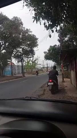 The video is taken from inside a vehicle looking out at a road. A person on a motorcycle is parked on the side of the road. Trees and some buildings are visible. The sky is overcast.