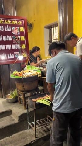 The video shows a street food vendor in Vietnam. Three people are visible in the foreground. Two men are grilling skewers over a small charcoal grill. The skewers appear to have some sort of meat or sausage attached to lemongrass stalks. One man is wearing a blue shirt and brown pants and is actively placing skewers on the grill. The other man is wearing a white shirt and stands to the right, appearing to observe the grilling process. The back of a third person, who is wearing a black garment, is on the left side of the frame.  Behind the grill, a woman is preparing food at a counter. She is wearing a black shirt and uses tongs to work with the ingredients. The food stall has a sign with prices written in Vietnamese, along with images of different dishes. Plates filled with colorful skewers and ingredients are displayed. The background is a vibrant yellow wall with a window. Decorative lanterns are hanging above the street. The setting appears to be an evening street scene.