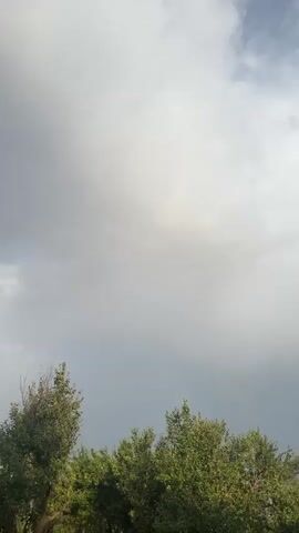 The video shows a time-lapse of clouds moving across the sky. The sky is mostly overcast, with some patches of blue sky visible. There are trees in the foreground, and a faint rainbow is visible in the distance. The video appears to have been filmed during the day.