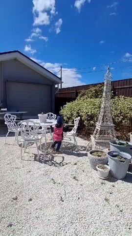 The video shows a small child walking in a backyard on a sunny day. The child, wearing a blue hat and a pink vest, is seen walking around a white metal table and chairs set. In the background, there is a white Eiffel Tower replica, along with several potted plants. The backyard is covered in white gravel. A building and a wooden fence can be seen in the background under a blue sky with scattered clouds.