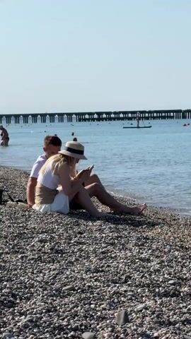A couple sits on a pebbled beach with the ocean in the background. The woman is wearing a white dress and a straw hat and is looking at her phone. The man is sitting behind her, with his arm around her. The water is calm and a pier extends into the distance. There are a few other people visible in the water.
