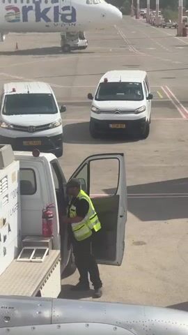 An Arkia airplane is parked on an tarmac. A white ground vehicle is positioned next to the airplane, and a man wearing a yellow safety vest is standing next to it with the door open. Two white vans are parked in the background.