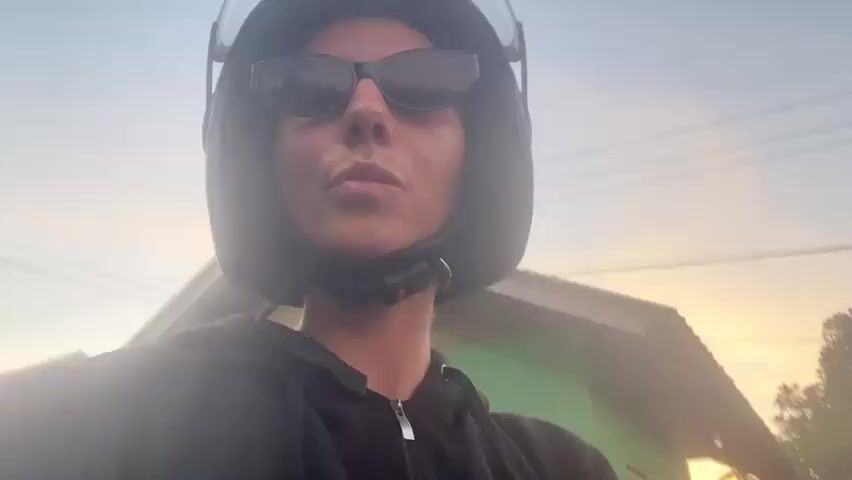 A woman wearing a black hoodie, sunglasses, and a blue and black helmet is riding on the back of a motorcycle. She is looking at the camera and making kissy faces. Behind her, a person wearing a gray t-shirt and a black helmet is riding the motorcycle and looking at their phone. The background is a rural landscape with trees, green fields, and a pink and orange sunset sky. The video appears to be taken during golden hour.