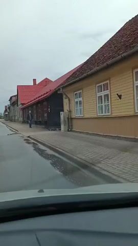 The video shows a view from inside a vehicle driving down a wet street lined with old wooden buildings with red roofs. A man in a dark jacket and jeans is walking on the sidewalk. The sky is overcast.