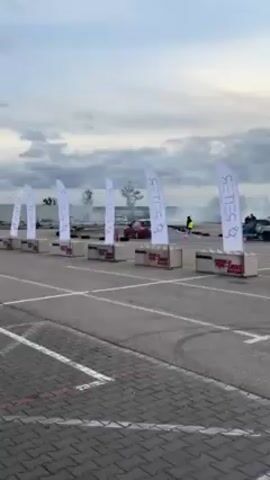 The video shows a red car drifting in a parking lot, with white banners and concrete blocks lining the edge of the track. The banners have the word "GETEC" written on them, along with a logo. The car is surrounded by smoke, and there are people wearing yellow vests standing around the perimeter of the track. The sky is cloudy, and the ground is paved with gray asphalt.
