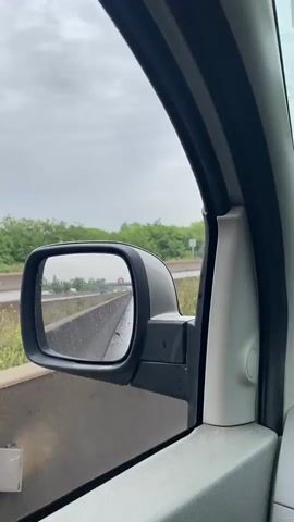 The view from inside a car looking out a side view mirror on a rainy day. The mirror is black and reflects the highway behind the vehicle. Other cars can be seen in the mirror, along with a speed limit sign. The highway is wet from the rain. Outside of the mirror, there is a barrier next to the road, followed by green trees and a cloudy sky.