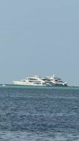 The video shows a large, white yacht on the open sea, viewed from a lower vantage point. The yacht has multiple decks and is situated in the distance, with a clear blue sky above. The sea transitions from a deeper blue in the foreground to a lighter, turquoise shade closer to the yacht, possibly indicating shallower waters or a reef. There is gentle movement in the water.