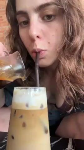 A person with long, wavy brown hair is shown from the chest up, holding a glass pitcher of dark liquid, presumably coffee. They are pouring the liquid into a tall glass filled with ice, coffee, and what appears to be a layer of foamed milk on top. The person is simultaneously drinking from the glass through a straw. The background appears to be a café setting, with a brick wall partially visible in the distance. A black and white marble-patterned table is visible at the bottom. The person is wearing a dark shirt, and a hint of a tattoo is visible on their chest. The lighting is bright and appears to be natural daylight.