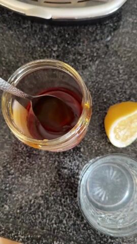 A high-angle close-up video shows a person preparing a drink. A glass jar is visible containing a thick, amber liquid. Next to the jar is a cut lemon half, and a clear glass of water with a cut design around the bottom. The person takes a spoonful of the amber liquid from the jar and drops it into the water glass. The spoon is then used to stir the liquid in the glass of water. The surface of the counter is a gray-colored, speckled material. The top of a stainless-steel appliance is partially visible in the background. There is the faint sound of a baby vocalizing.