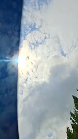 A shot of the sun shining through clouds. The sun is on the left side of the frame, and the clouds fill the rest of the screen. A green tree is visible on the right side of the frame.