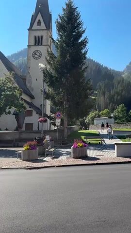 The video pans across a picturesque village scene on a clear, sunny day. In the foreground, a paved road runs horizontally. To the left, a church tower with a black spire and a clock face dominates the view, partially obscured by a tall, dark green coniferous tree. Flower boxes filled with colorful blossoms are placed near benches in a paved public area. Several people are walking in the background. Further to the right, a white house with a red-tiled roof is visible, with a white car parked in its driveway. Beyond the house, a densely wooded mountain slopes upwards, with a ski slope visible on its upper half. The sky is a bright, clear blue, and the overall atmosphere is tranquil and idyllic.