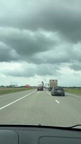 The video depicts a view from inside a car driving on a multi-lane highway on a cloudy day. Several vehicles are ahead, including a tanker truck, a box truck, and a dark grey car with a noticeable quad exhaust system. The dark grey car is in the same lane and slightly ahead of the recording car. The landscape on either side of the road consists of flat, grassy fields. Road signs are visible along the side of the road. The sky is overcast with dark, heavy clouds.